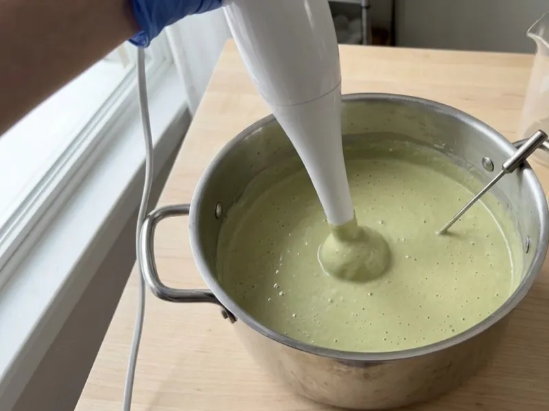 A gloved hand blends pale green soap mixture in a stainless steel pot using an immersion blender.