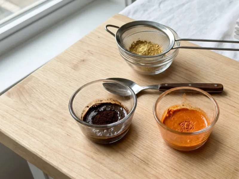 Natural soap colorants in small glass bowls, a strainer, spoon, and oil on a wooden board.