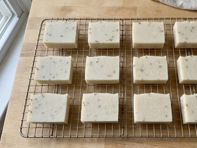 Twelve bars of off-white handmade soap with dried herbs curing on a wire rack.