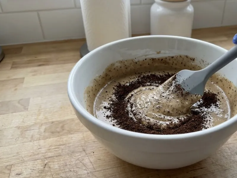 A gloved hand stirs coffee grounds into creamy soap batter inside a white mixing bowl.