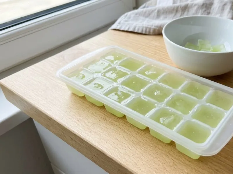 Light green liquid frozen in an ice cube tray on a wooden counter beside a small bowl.