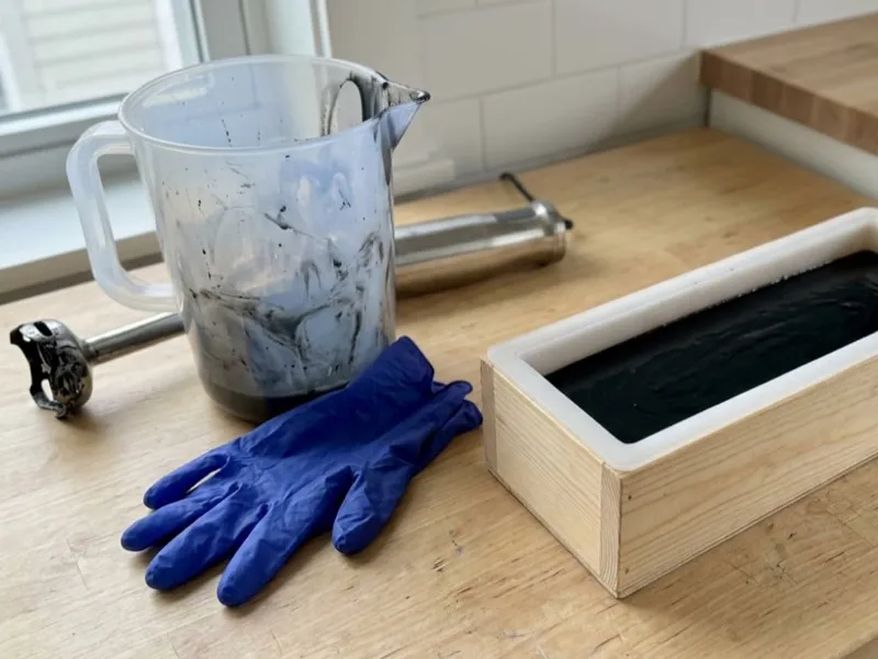 Black soap batter rests in a loaf mold beside a mixing pitcher, immersion blender, and blue gloves.