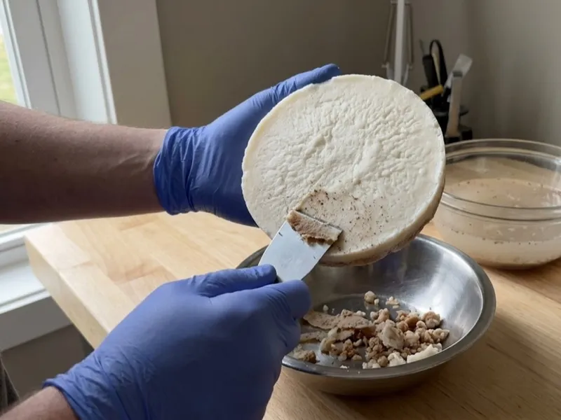 Gloved hands scrape impurities from a round disc of rendered tallow into a metal bowl.