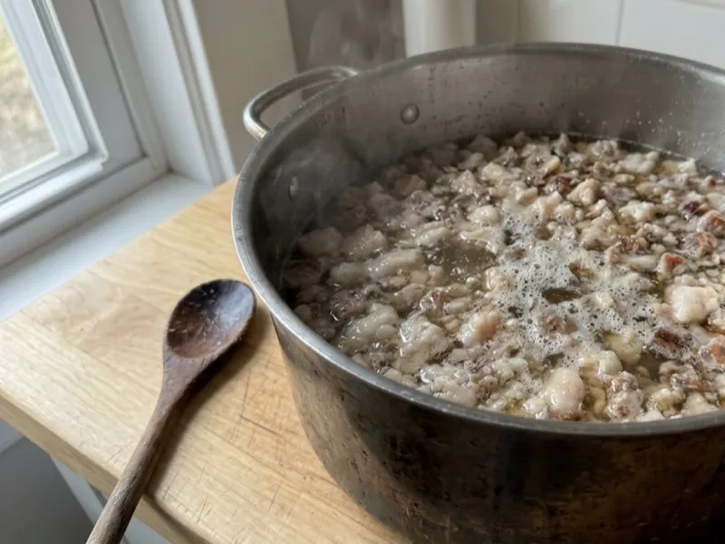 Small pieces of animal fat rendering in a stainless steel pot next to a wooden spoon.